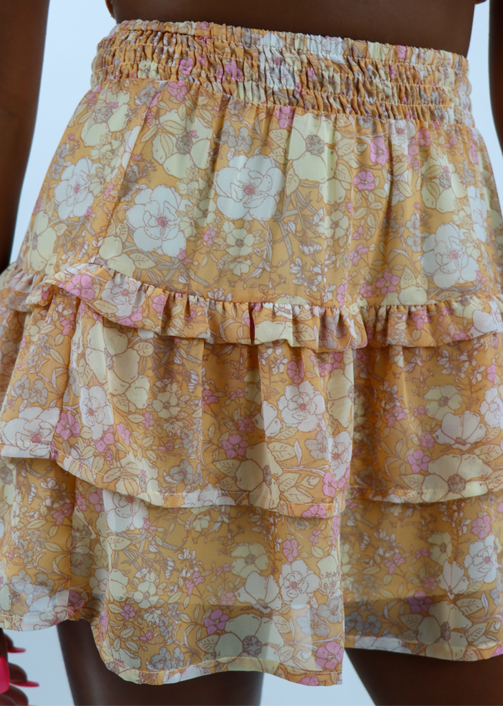 Orange skirt with white and yellow flowers. Features a smocked elastic waistband with tiered ruffle detailing. This is the skirt portion of a two-piece set